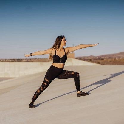 A female model showcasing a black WILDRAX Balconette Sports Bra while striking a warrior yoga pose on a concrete structure outdoors.