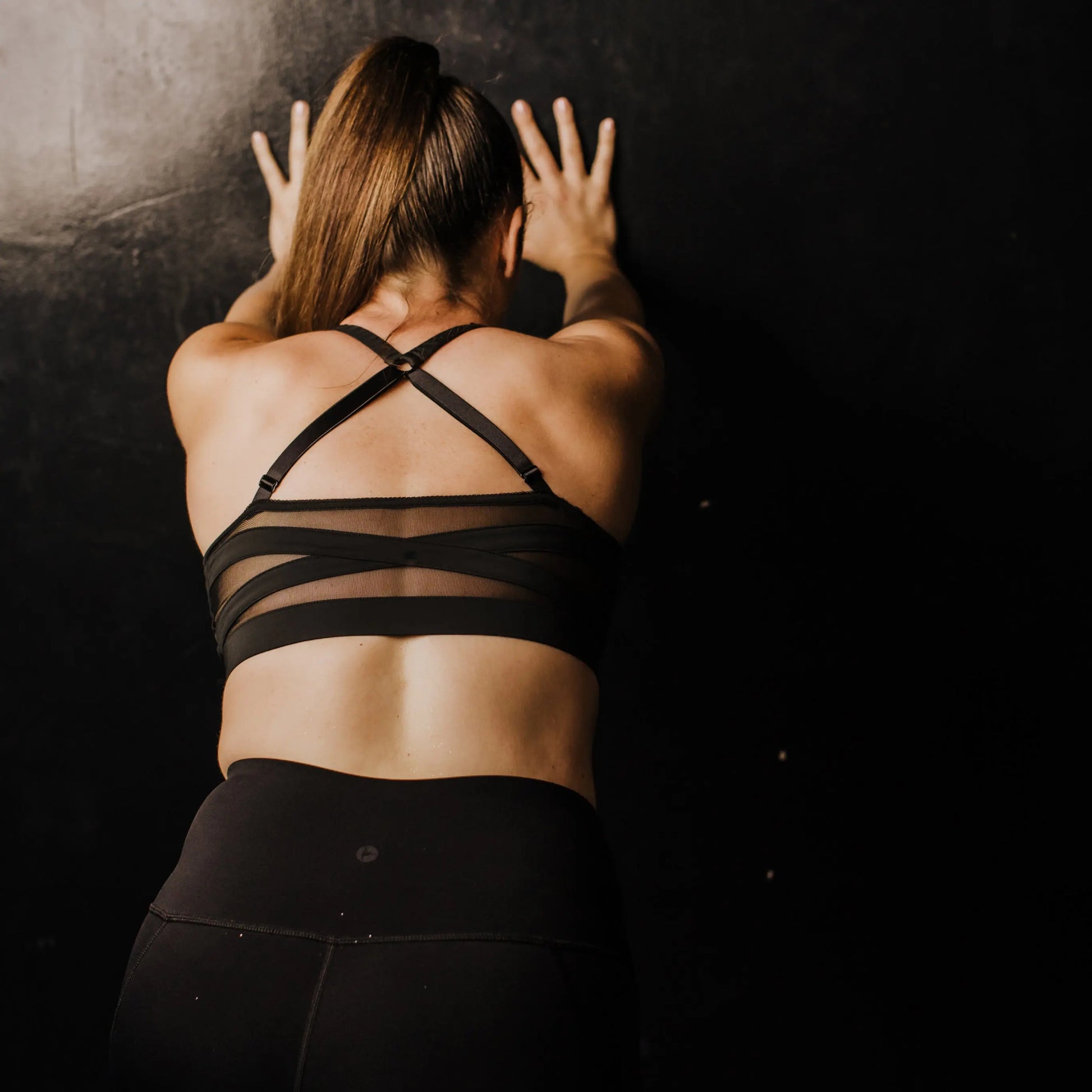 A woman showcases the ultimate comfort and fit of the black power mesh back panel for Wildrax Balconette Sports Bra. The secure hold, achieved through the elastic band and adjustable racer back straps, is proudly displayed on the back of the model as she presses her hands against a black wall